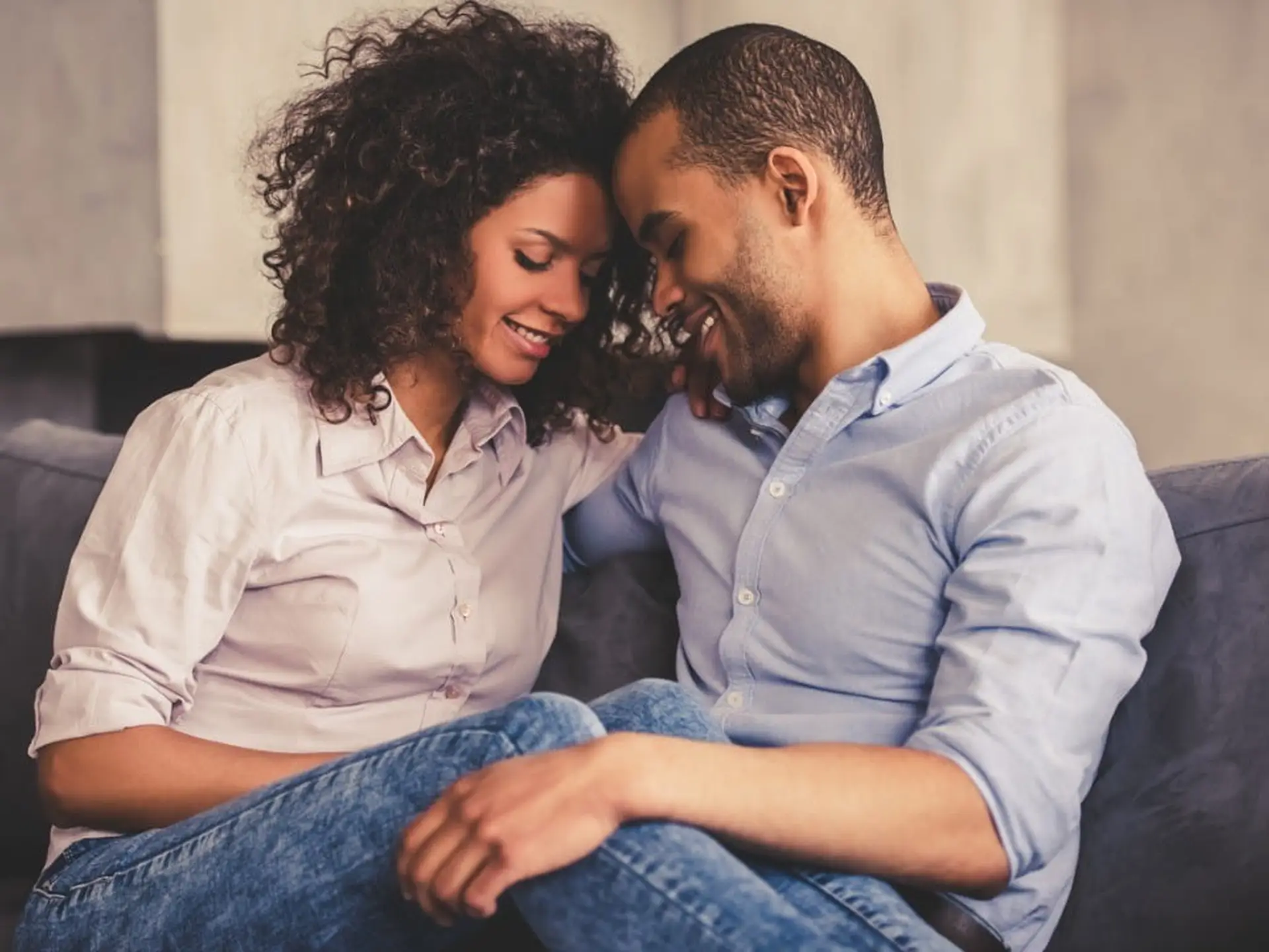 A couple sitting closely on a couch, smiling and leaning their heads together, both wearing casual shirts and jeans.