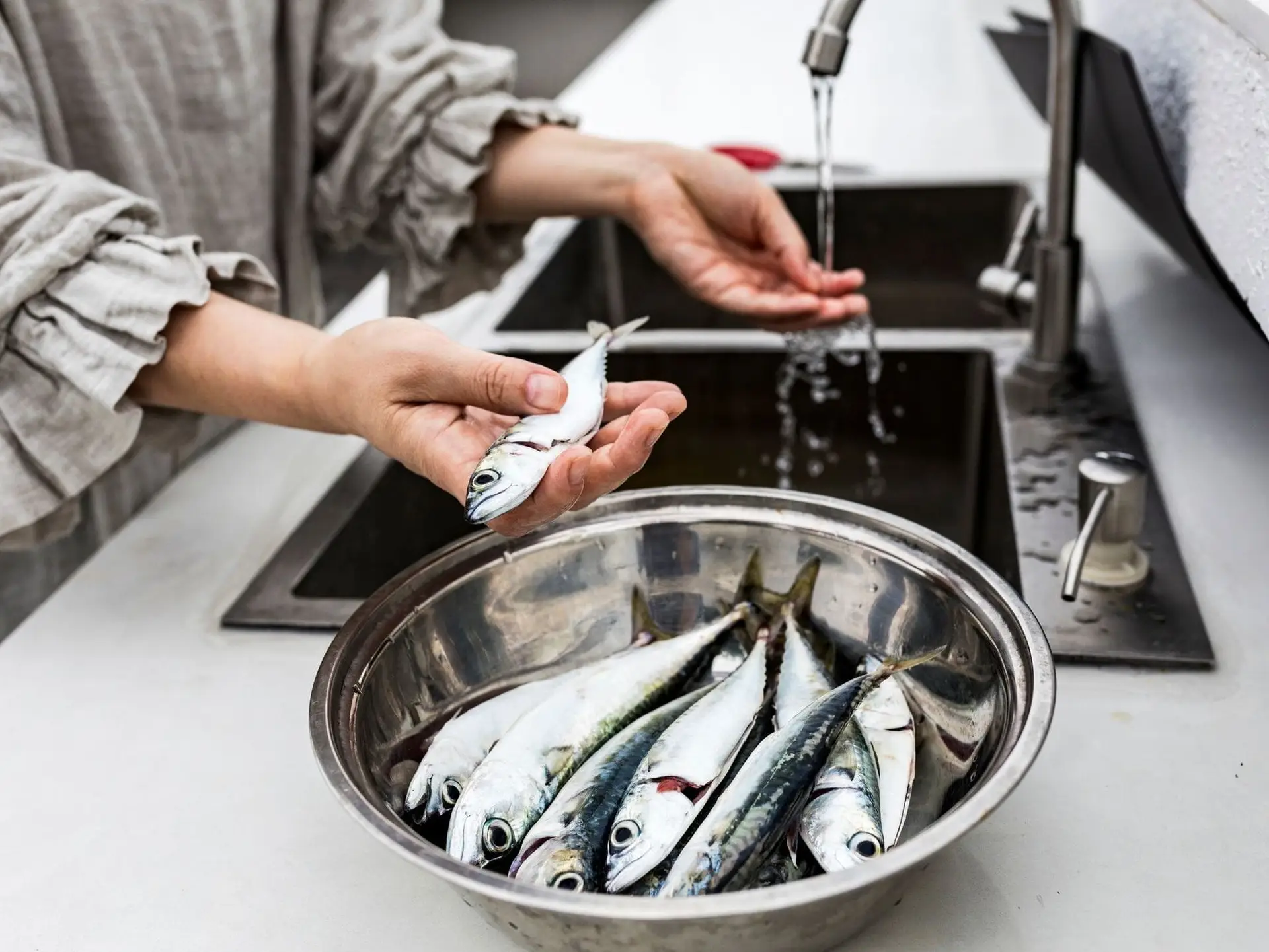 Person rinsing small fish at a sink, holding one in hand, with more fish in a metal bowl on the counter.