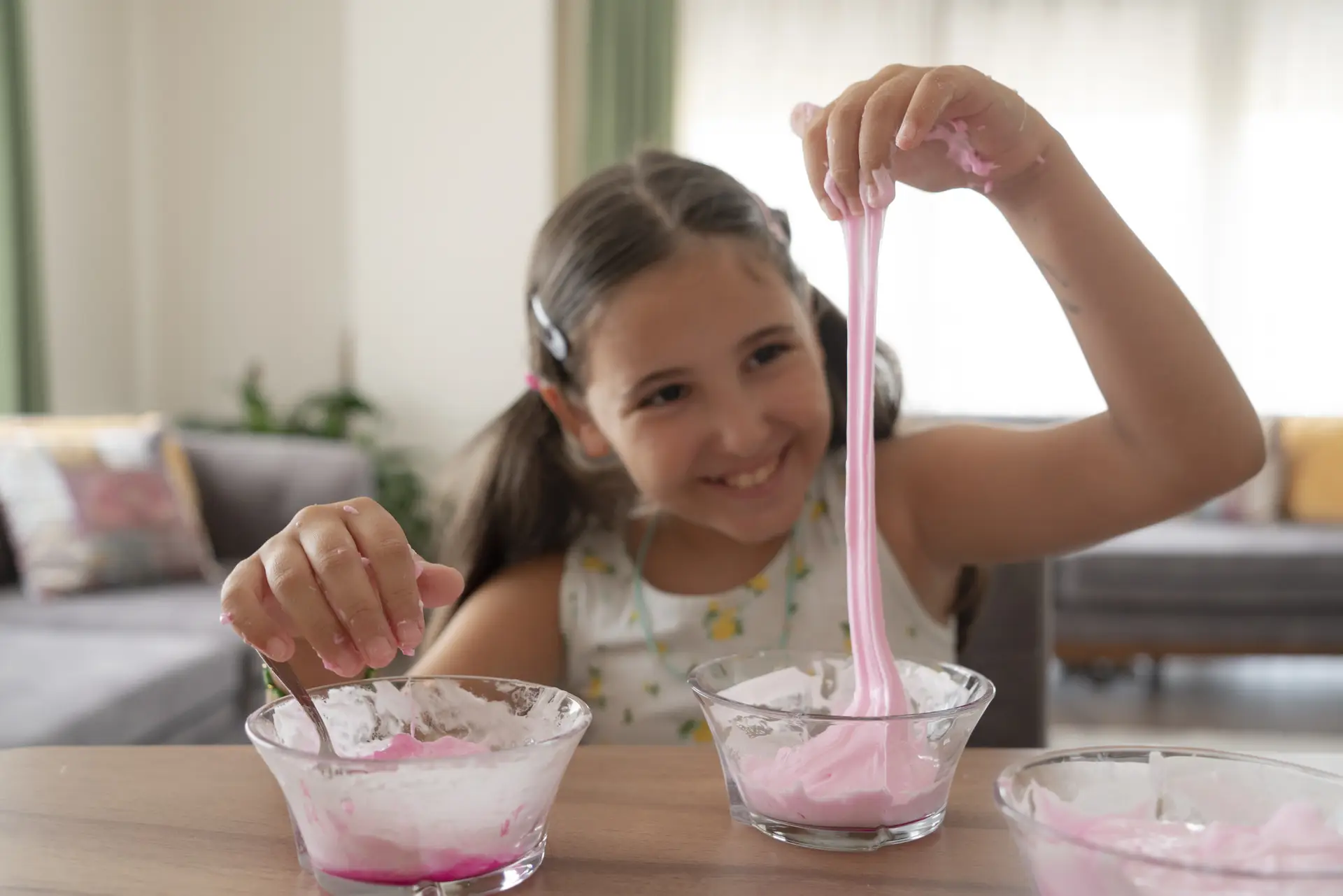 A smiling young girl playing with pink slime in a bright living room, with bowls of slime on a wooden table.