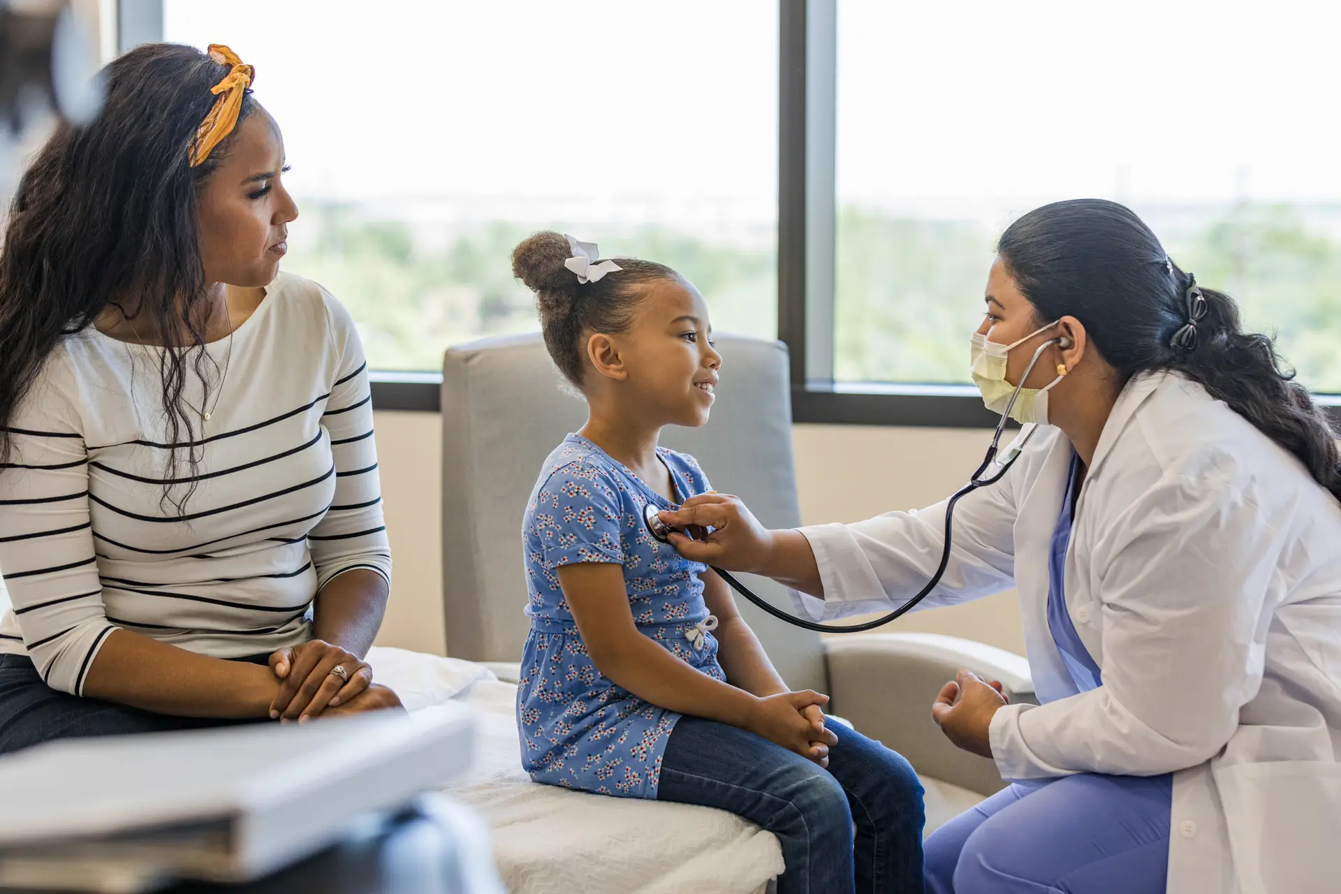 A doctor listens to a young girl's heartbeat with a stethoscope while her mother watches. They are in a brightly lit medical office.