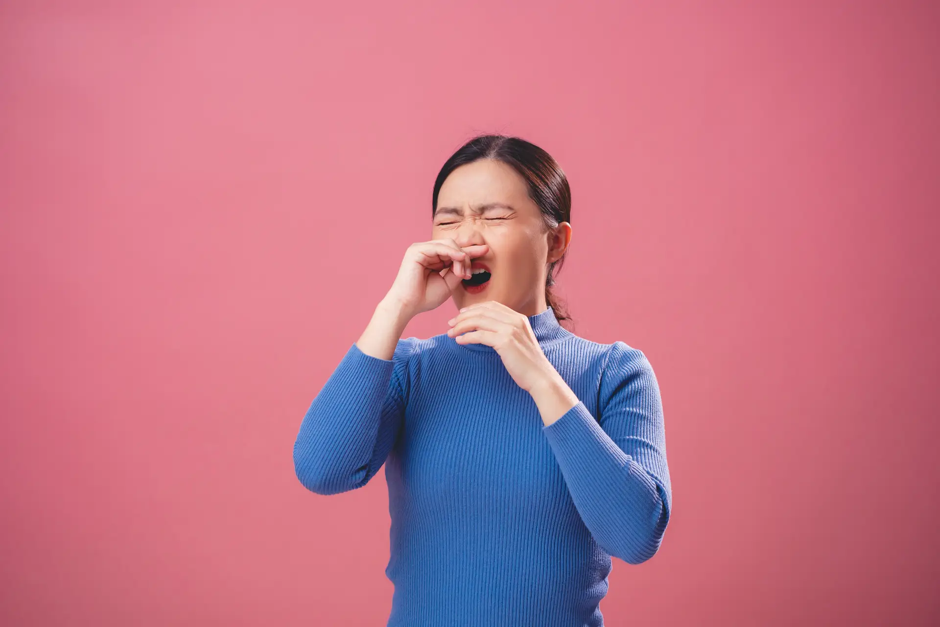 Person in a blue sweater sneezing into their hand against a pink background.