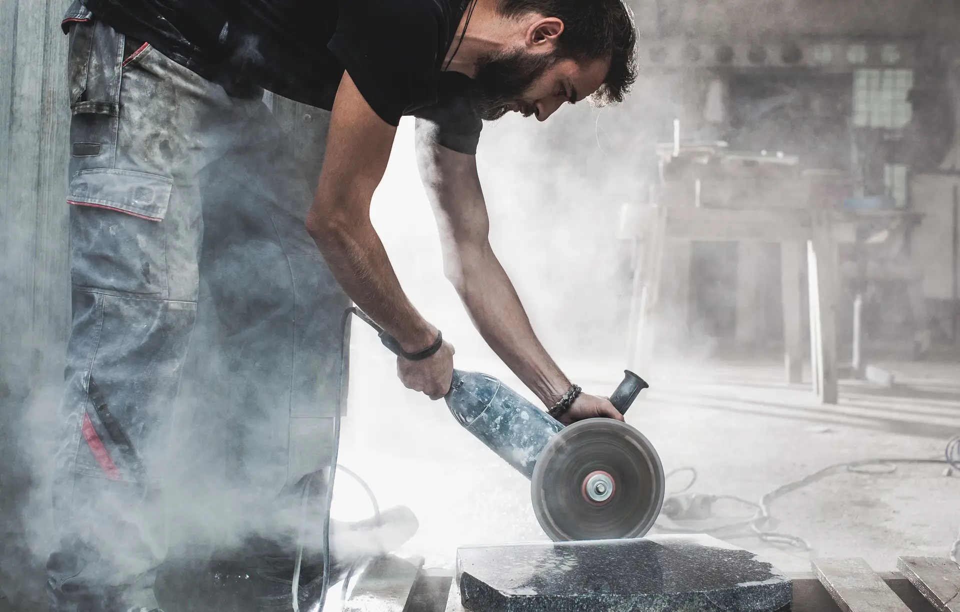 A man using an angle grinder to cut a stone block in a workshop filled with dust.