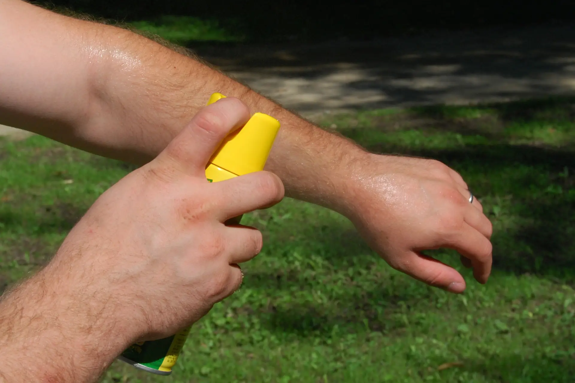 Person spraying insect repellent on their forearm outdoors, with a grassy background.