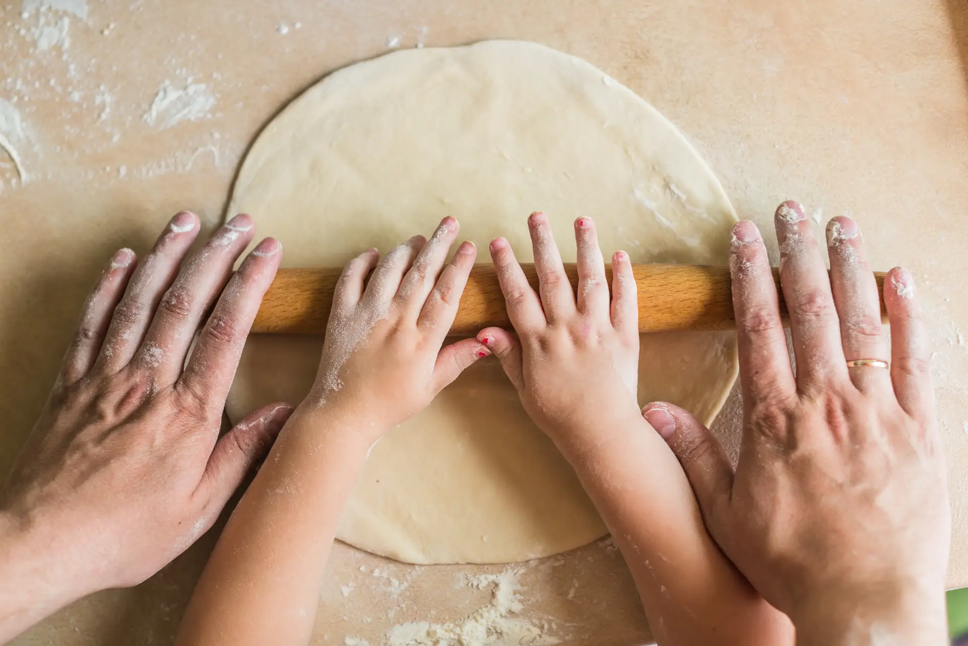 Adult and child's hands rolling dough with a wooden pin on a flour-dusted surface.