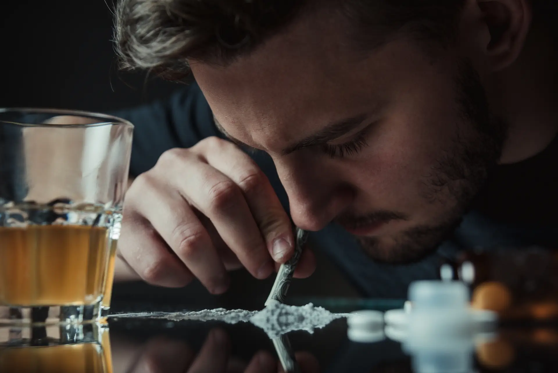 A person snorting a powdered substance through a rolled bill on a mirrored surface, with a glass of whiskey and pills nearby.