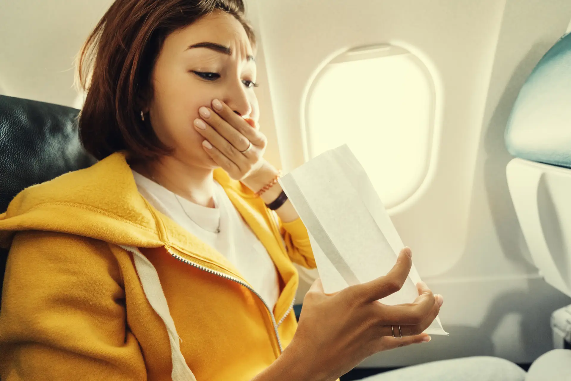 A woman in a yellow hoodie looks worried on an airplane, holding a sickness bag with one hand and covering her mouth with the other.