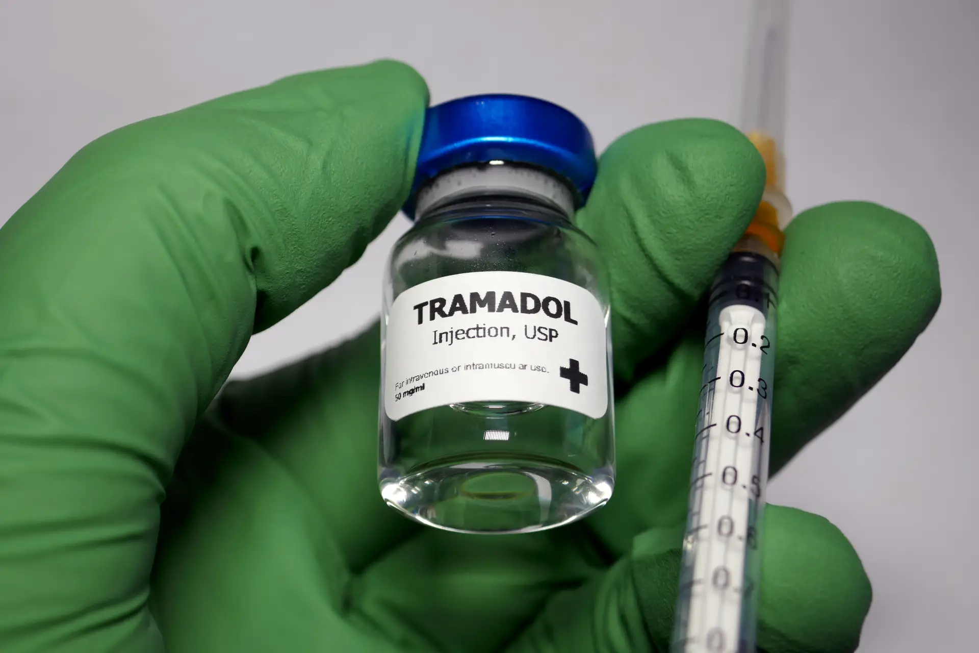 A green-gloved hand holds a vial labeled "Tramadol Injection, USP" and a syringe with measurement markings.