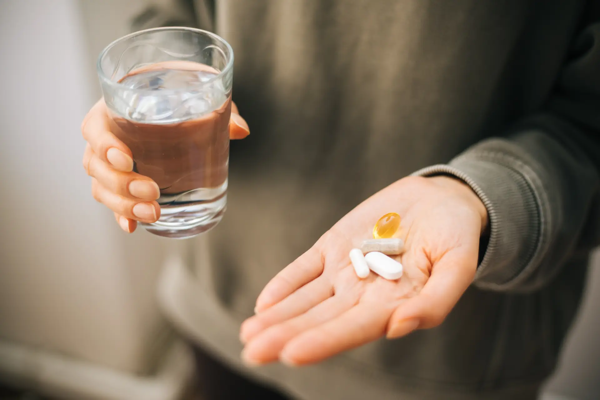 A person holding a glass of water in one hand and assorted pills in the other hand, wearing a green sweater.