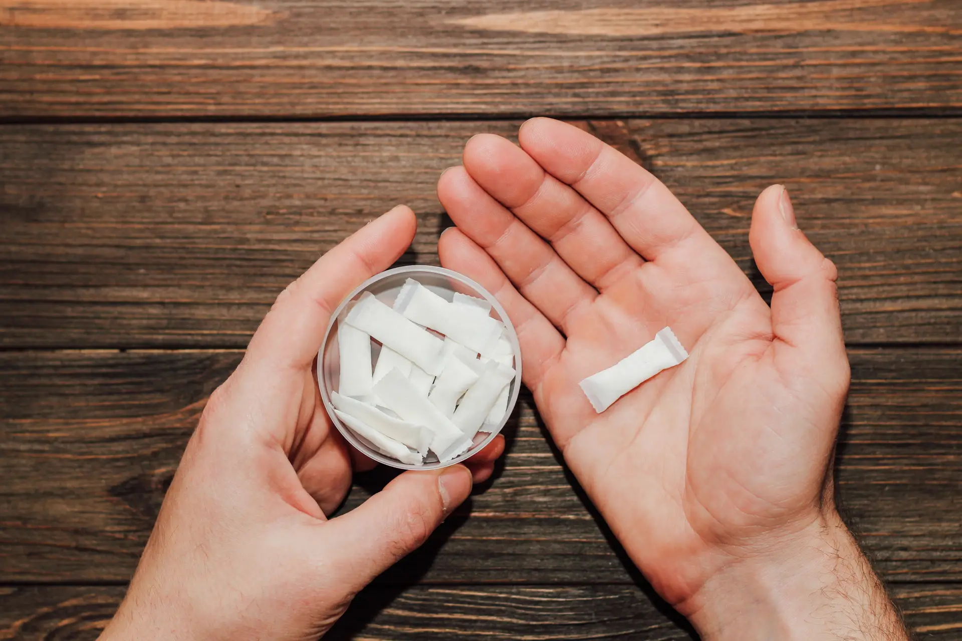 Hands holding a small container filled with white, rectangular packets and one packet in an open palm, on a wooden surface.