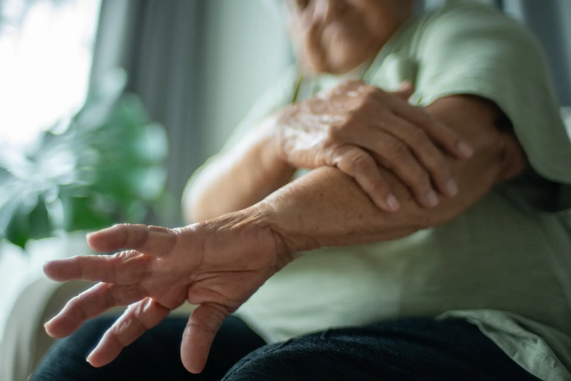 Elderly person in a green shirt rubs their own arm, possibly experiencing discomfort, with a blurred background of indoor plants.