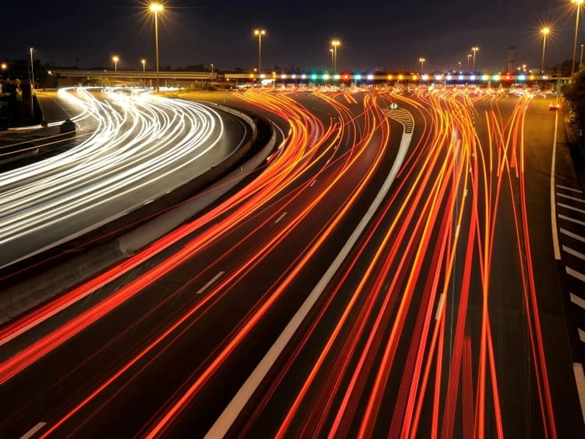 Long-exposure photo of a highway at night, showing red and white light trails from vehicles, with overhead streetlights illuminating the scene.