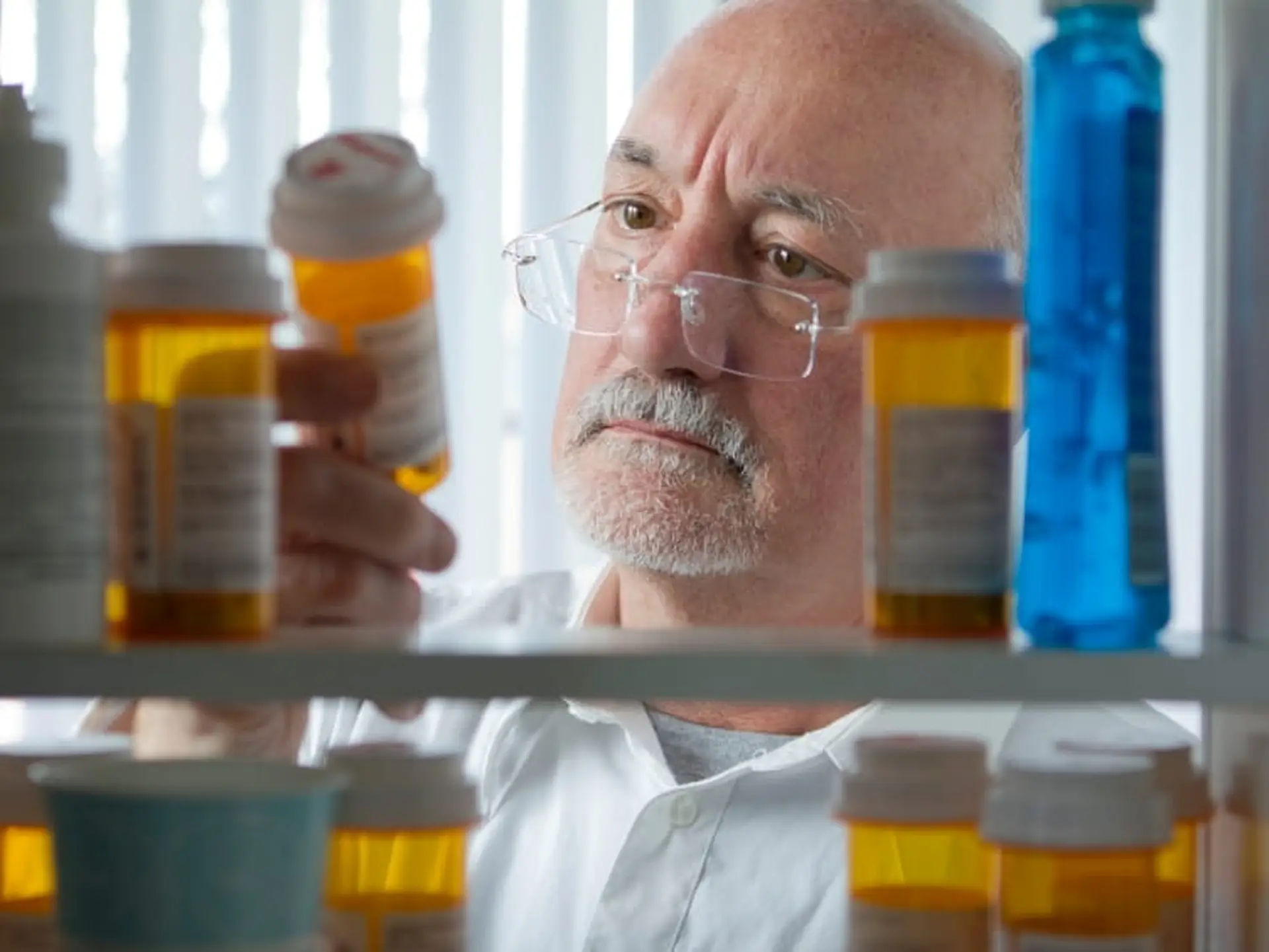Man with glasses examining a prescription bottle, surrounded by various medication bottles on shelves.