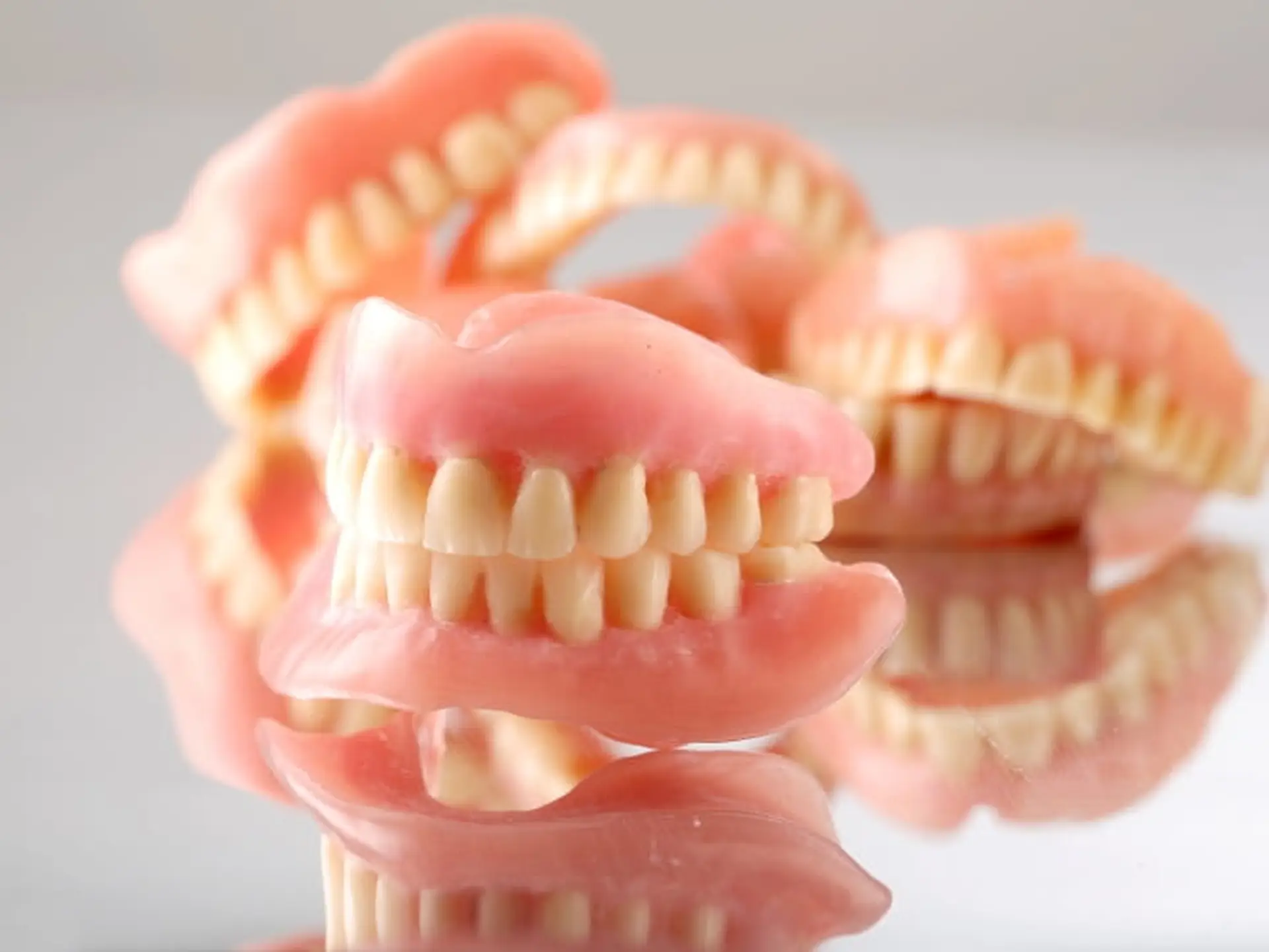 Several sets of artificial dentures with pink gums and white teeth are displayed on a reflective surface.